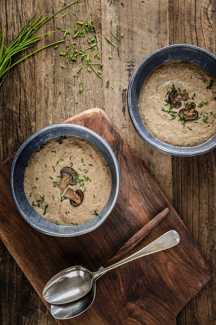 Two bowls of thick and creamy mushroom soup garnished with mushroom slices and chives in blue bowls on a wooden surface
