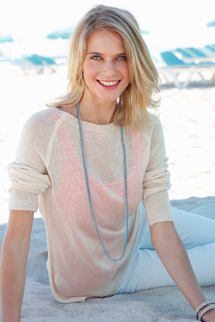 A blonde woman wearing a knitted jumper, white trousers and a long necklace at the beach