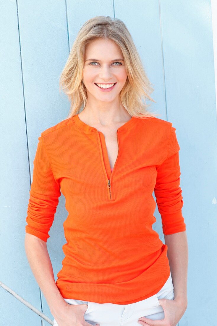 A blonde woman wearing an orange long-sleeved top with zip and white trousers