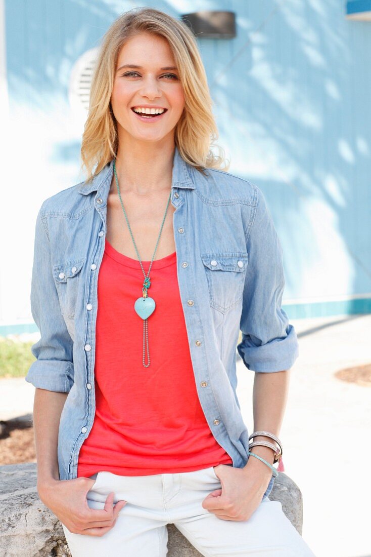 A blonde woman wearing a red top, denim shirt, white trousers and necklace