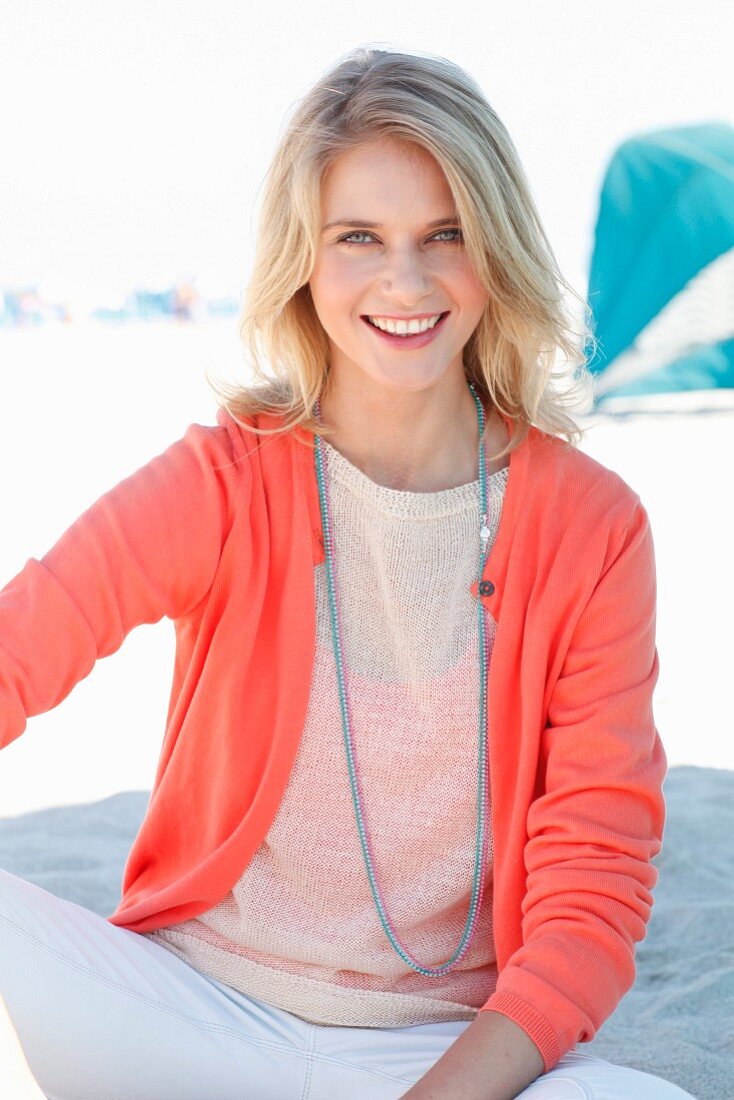 A blonde woman wearing a knitted jumper, a salmon-coloured cardigan and white trousers on the beach