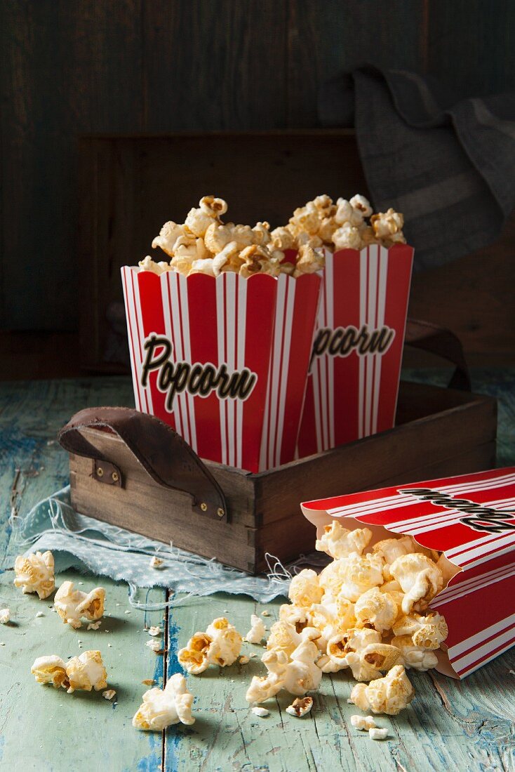 Two red and white striped boxes of popcorn on a wooden tray one box tipped over with popcorn spilling out onto an aqua green blue wooden surface