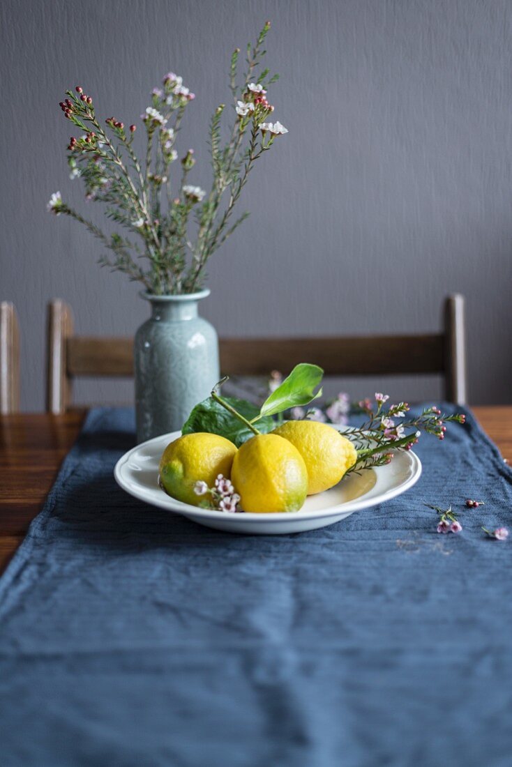 Zitronen auf weißem Teller vor Blumenvase auf Tisch