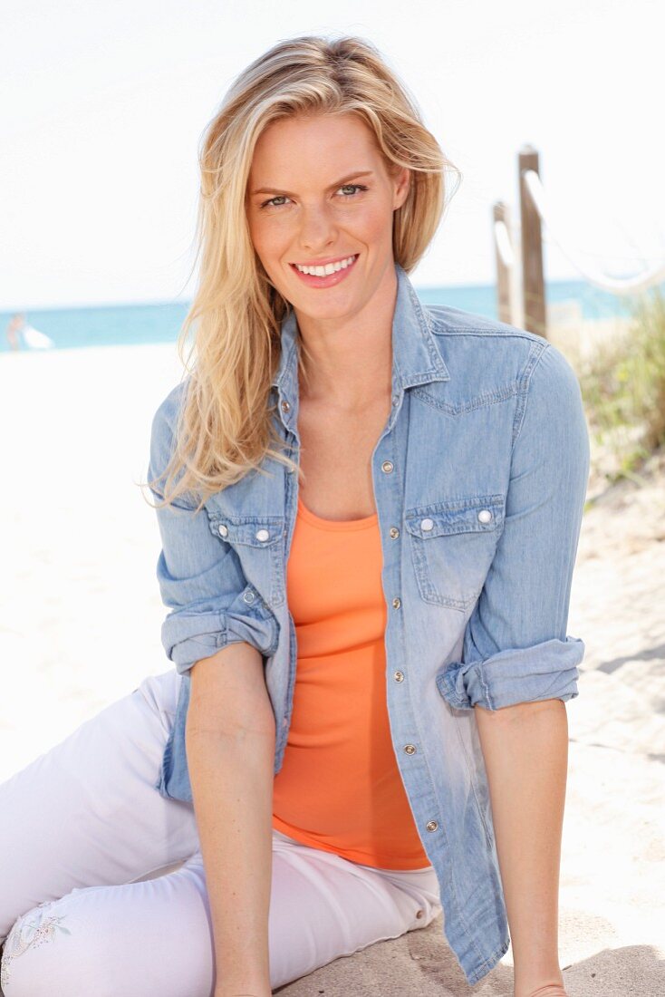 A blonde woman wearing an orange top, a denim shirt and white trousers on the beach