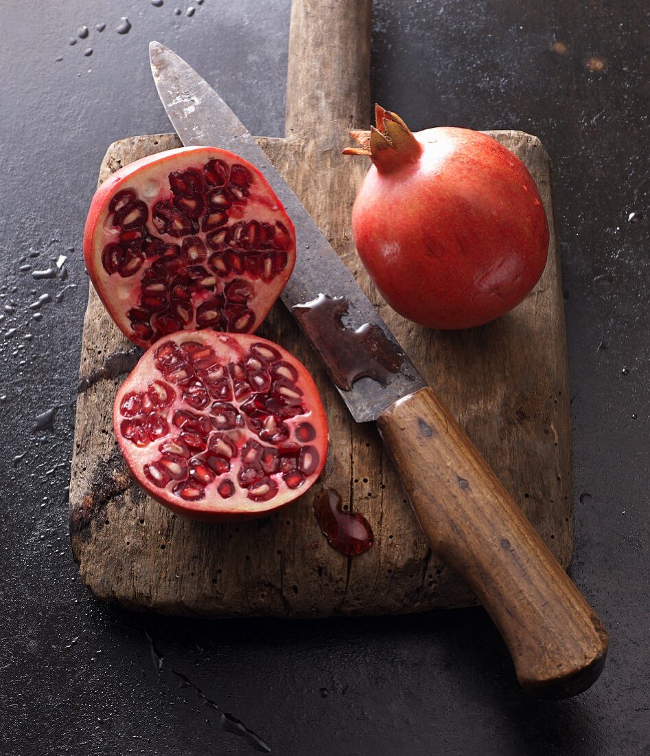 Two pomegranates, whole and halved, with a knife on an old wooden board