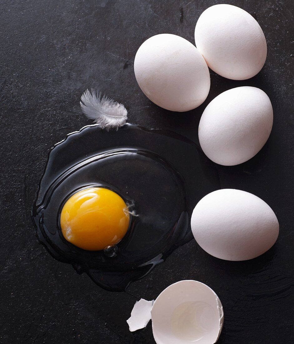 Whole eggs and egg yolk with a broken shell and feather on a black baking tray