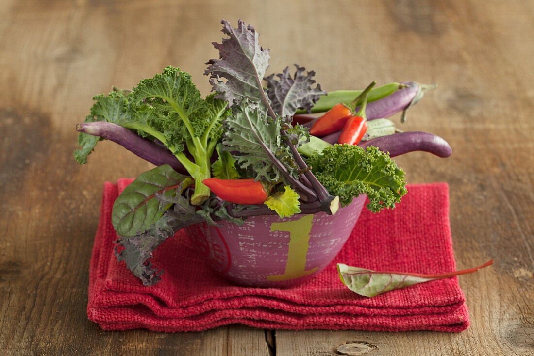 A vegetable bowl containing kale, chillis and long, thin aubergines