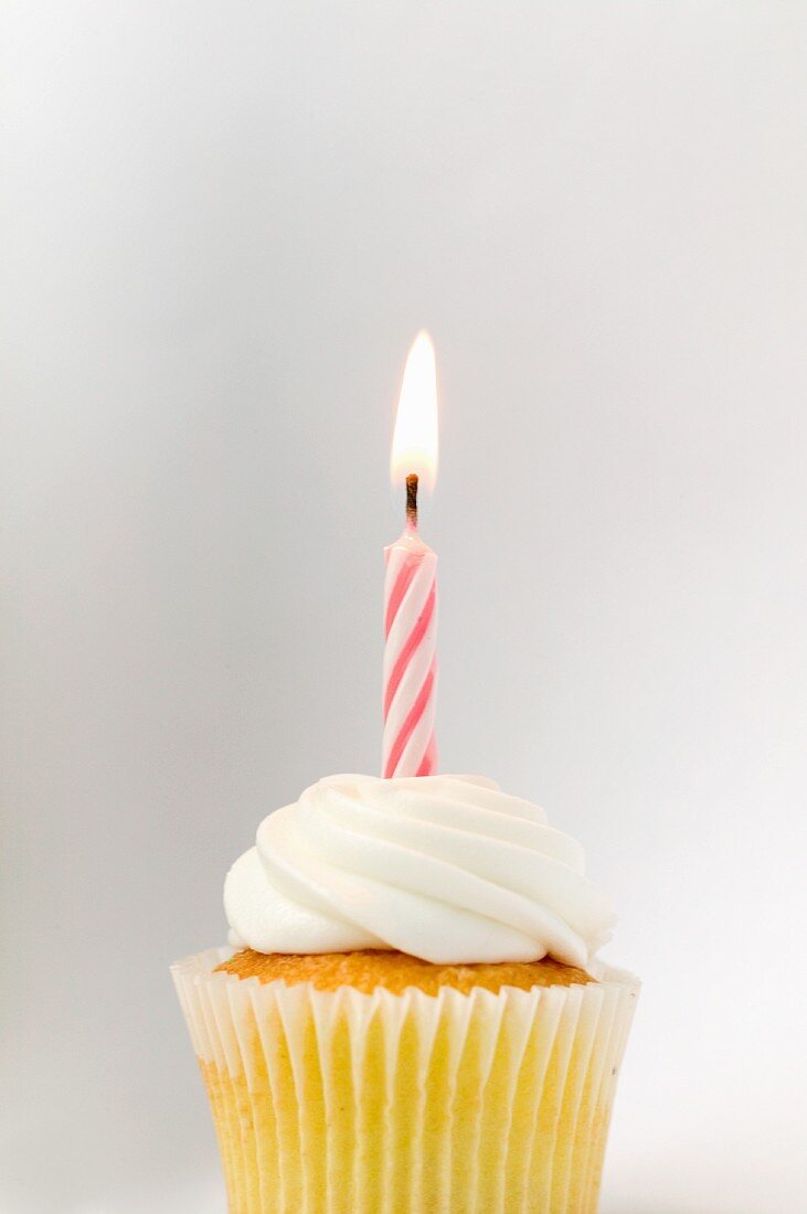 A birthday cupcake with a burning candle in front of a white background