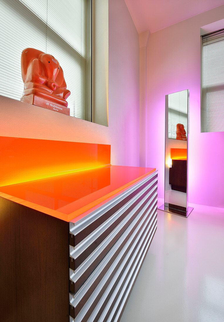 Indirect, orange and pink lighting around mirror and chest of drawers