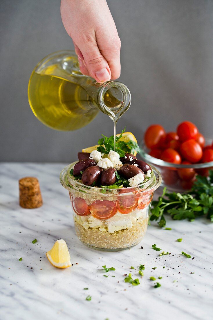 Olive oil being poured over a quinoa salad with feta, tomatoes and kalamata olives in a glass jar