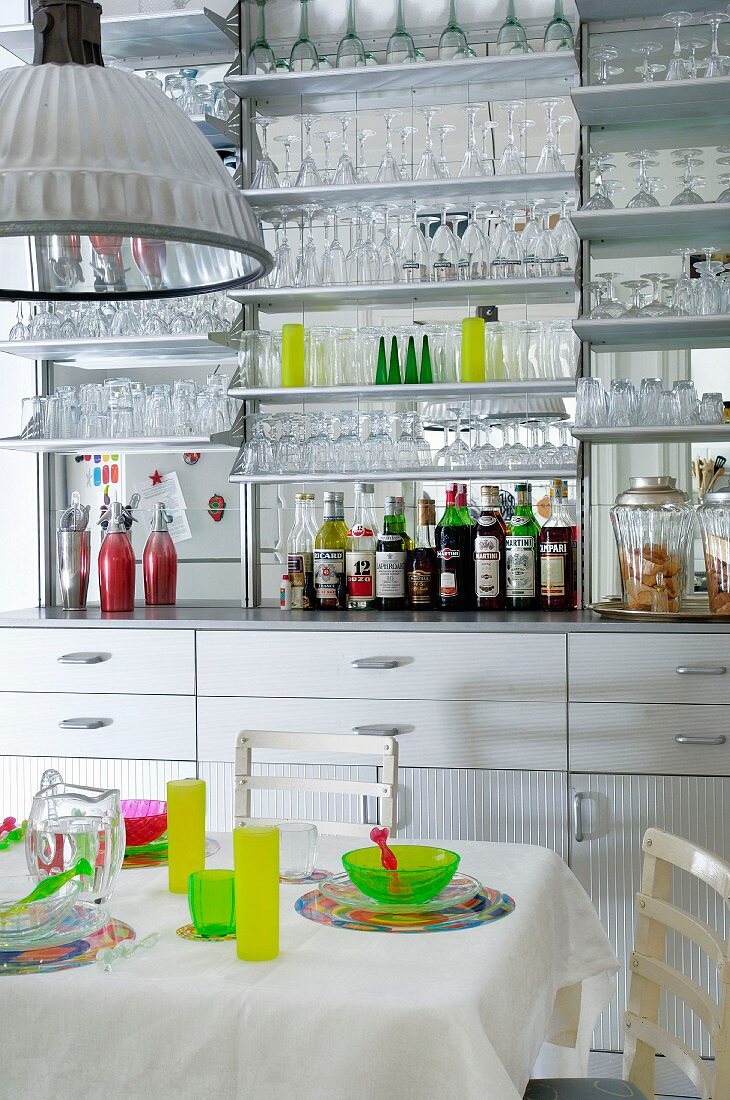 Kitchen counter with mirrored splashback and many glasses on wall-mounted shelves behind colourful place settings on white table