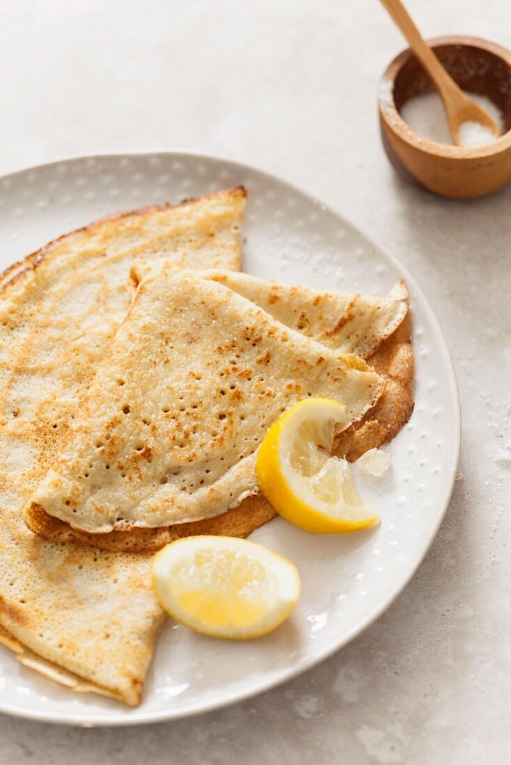 Two folded pancakes on an off white texture plate with lemon wedges on a stone surface