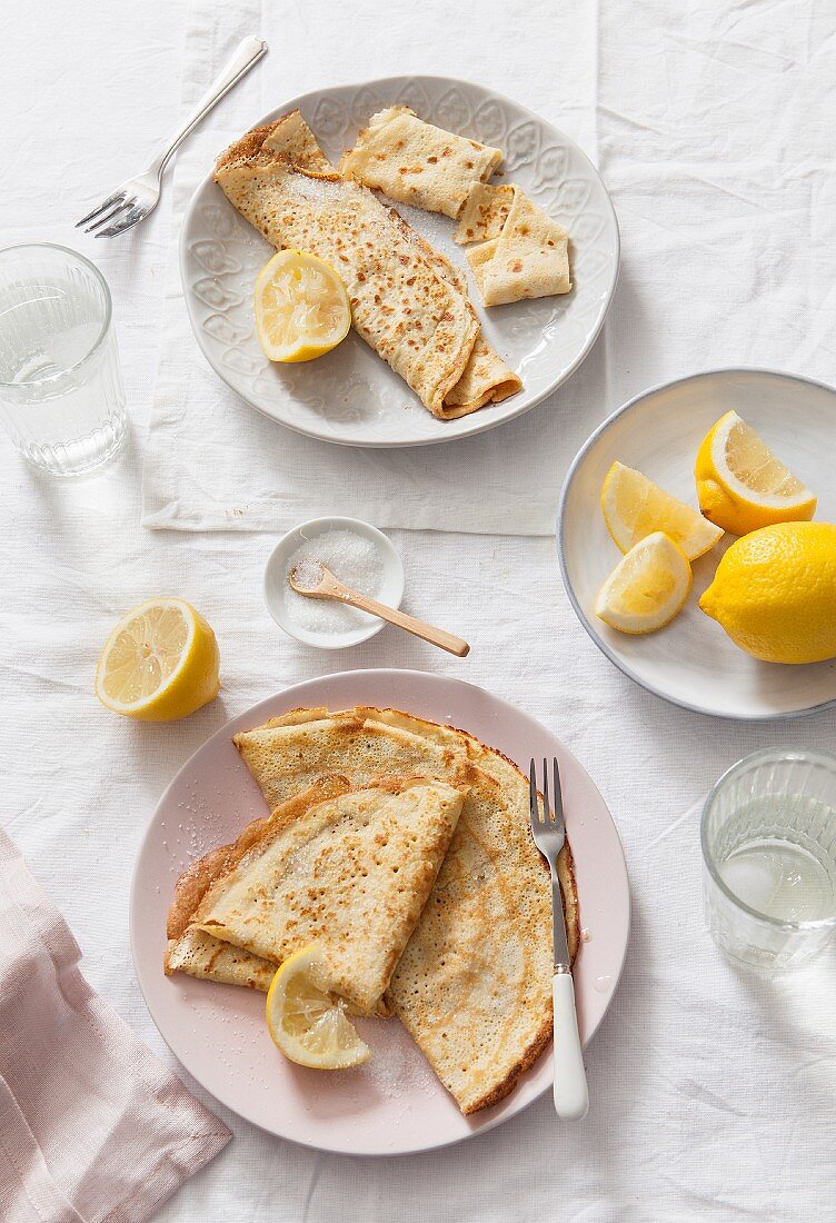 Two plates of folded and rolled pancakes served with lemon wedges on white linen covered table and water glasses