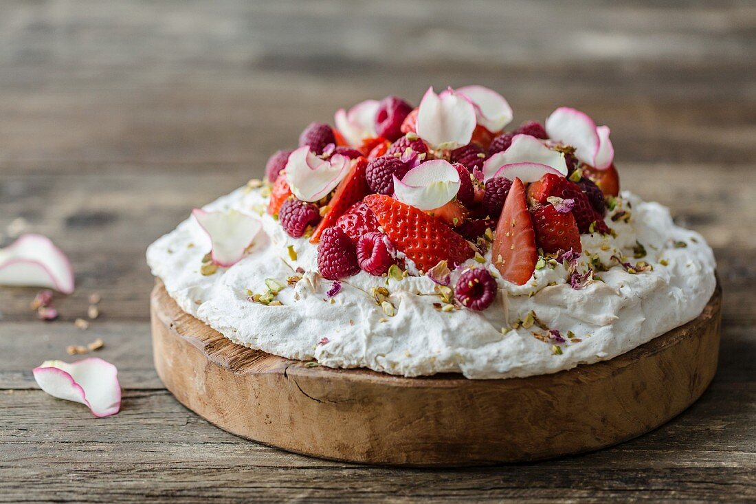 Rose and pistachio pavlova with fresh strawberries and raspberries, decorated with pistachio nuts and dried and fresh rose petals on a wooden surface