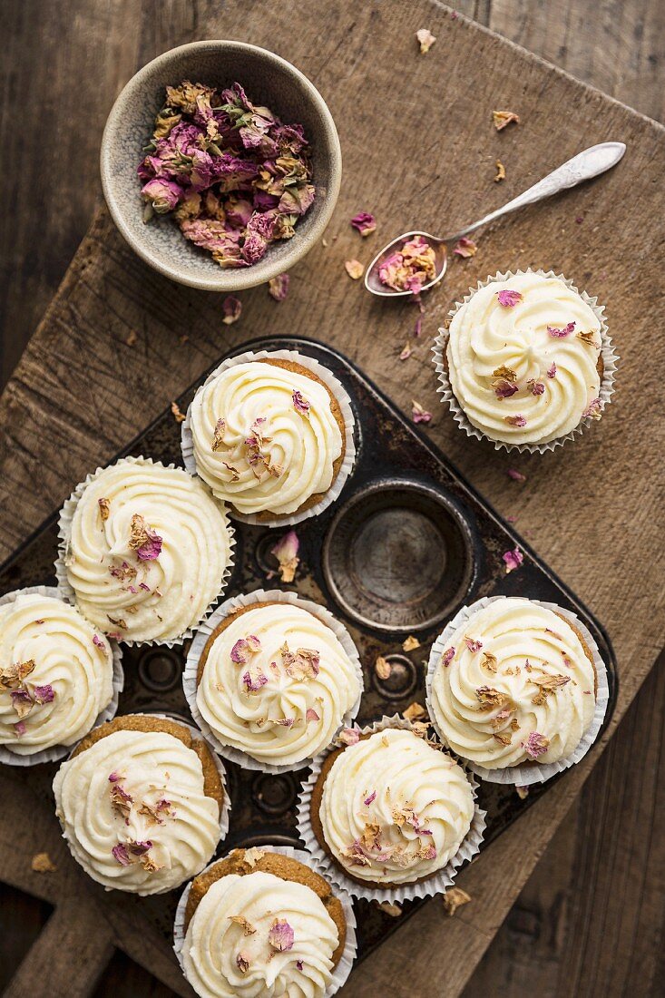 Rose and lemon cupcakes with rosewater and lemon frosting in a rustic baking tin decorated with dried edible rose petals with spoon and bowl of rose petals on a wooden board and table