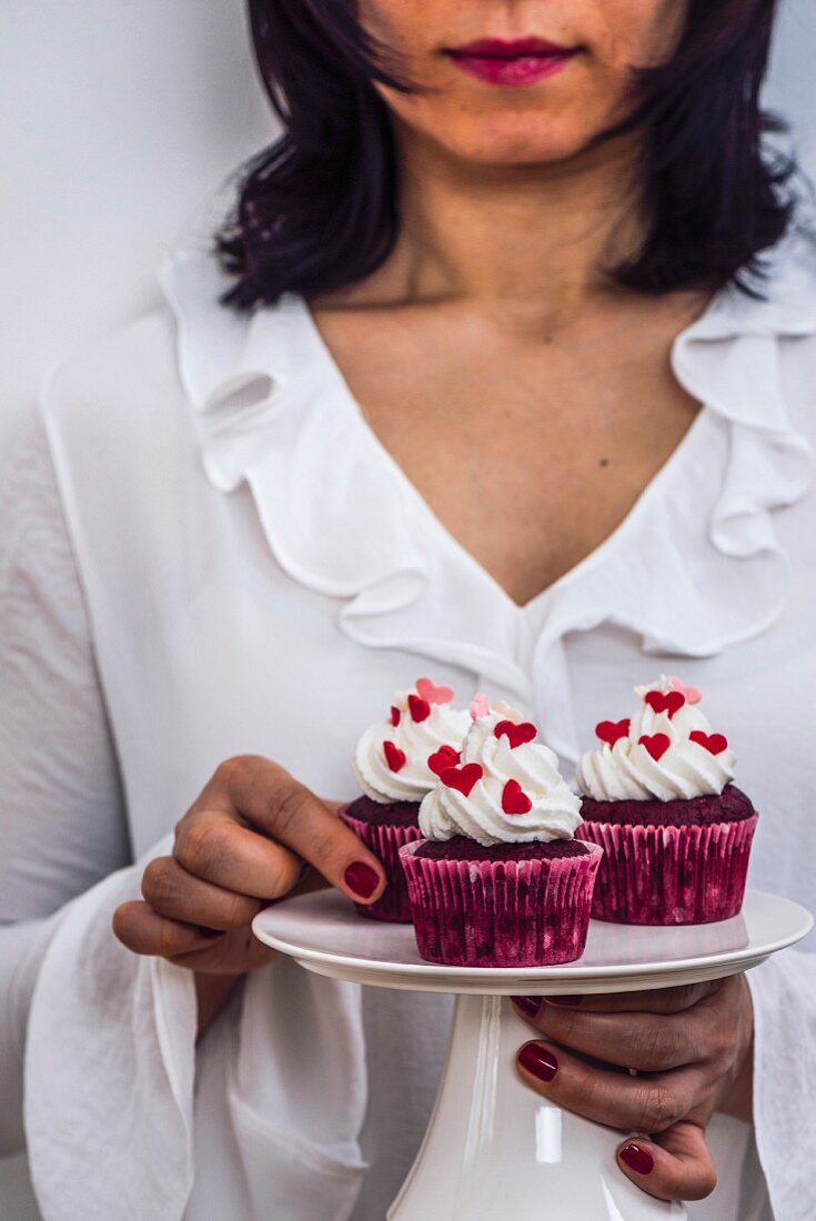 A woman with a white dress holding a cake stand full of red velvet cupcakes