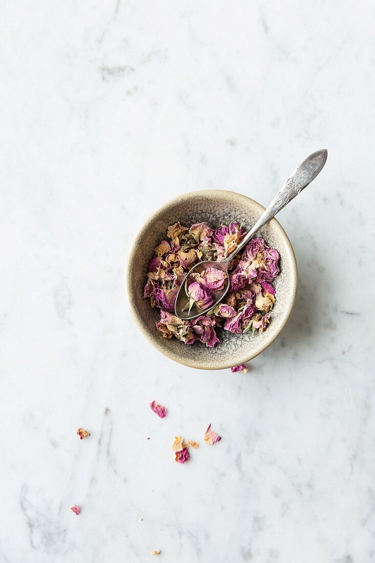 Bowl of dried edible pink rose petals and metal spoon on a grey and white marble surface
