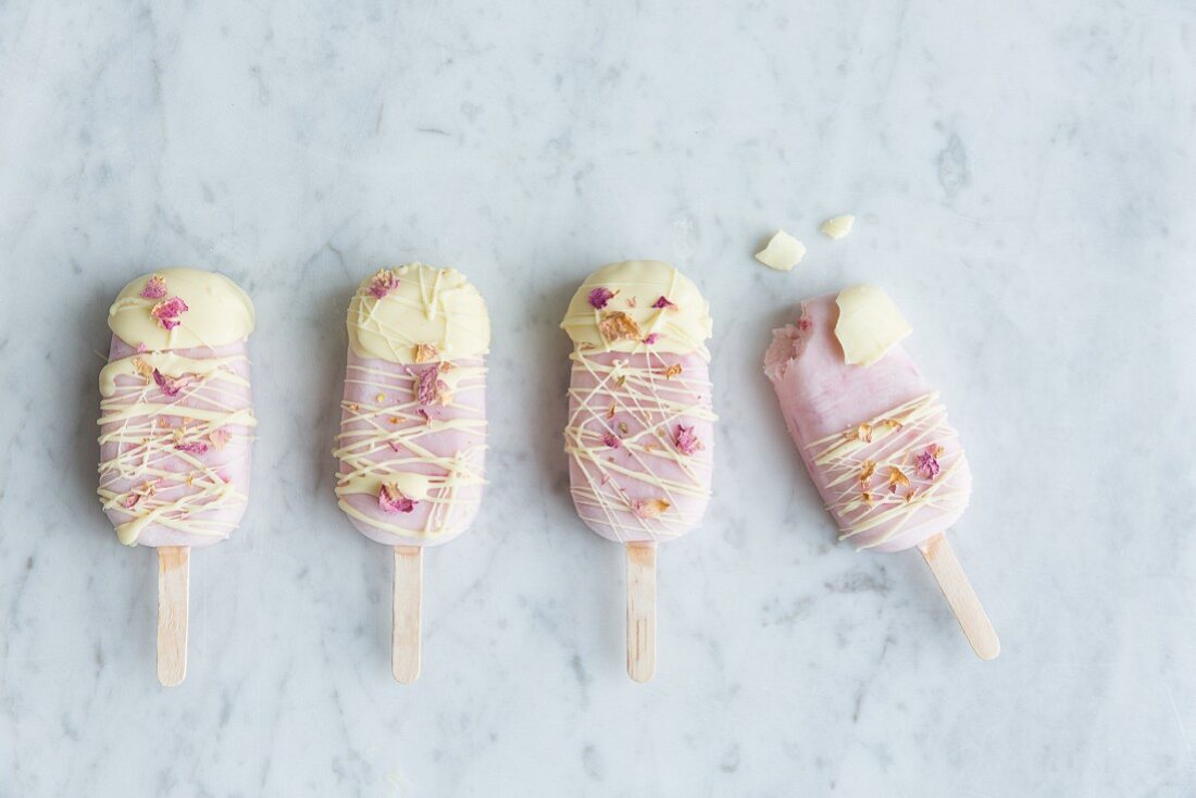 Rose and raspberry yoghurt frozen popsicles decorated with white chocolate and dried edible rose petals on a grey marble surface, one popsicle with a bite taken