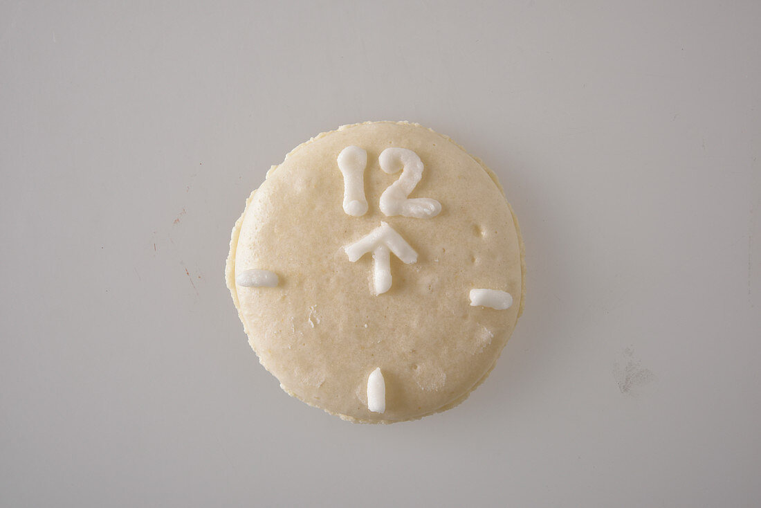 A white macaroon decorated to look like a clock with an arrow pointing to the 12