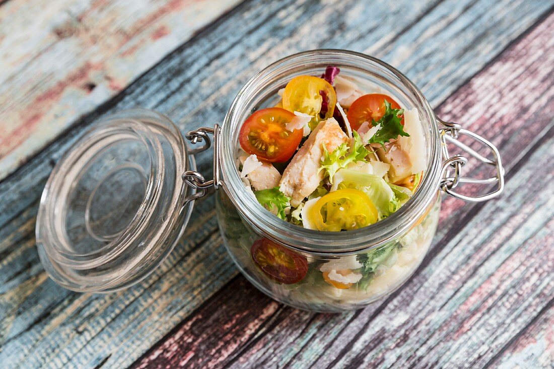 A pasta salad in a glass jar with tomatoes, chicken strips and pecorino