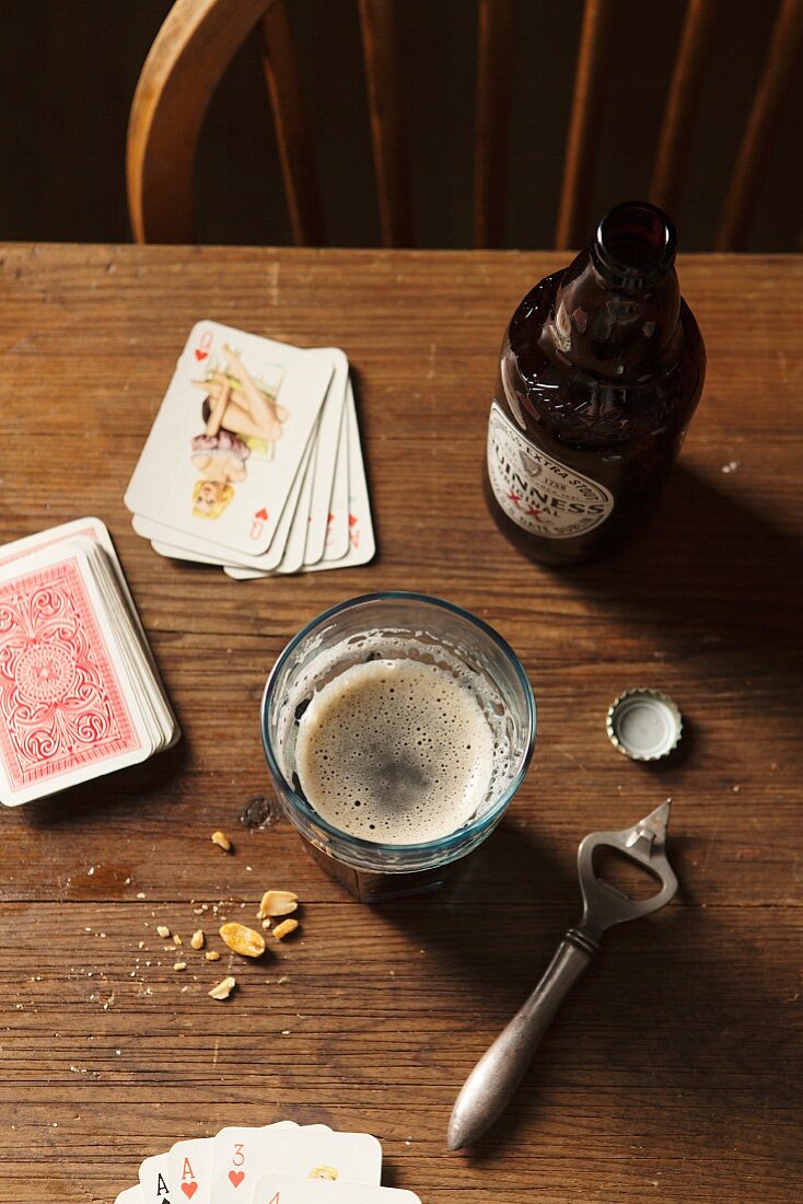 Guinness in Flasche und Glas, Flaschenöffner, Erdnüsse und Spielkarten auf Holztisch