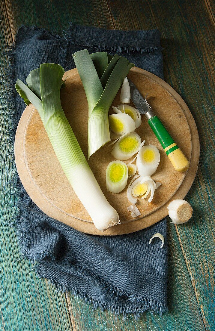 Overhead shot of two leeks on a round wooden chopping board being sliced by a retro wooden handled knife and navy cloth and green wooden background