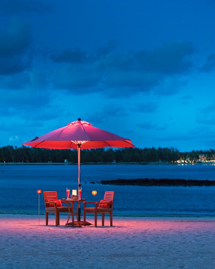 Romantische Abendstimmung an Sandstrand mit beleuchtetem Sonnenschirm und Armlehnstühlen