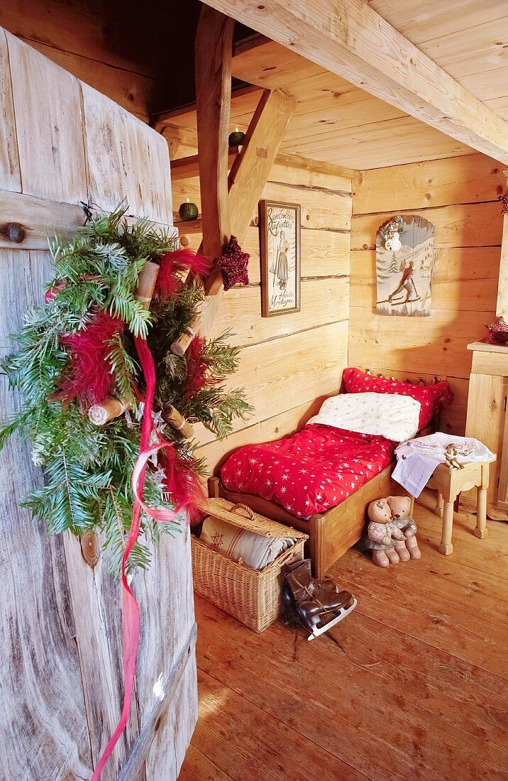 View of small bed in wooden Alpine cabin seen through open front door with wreath