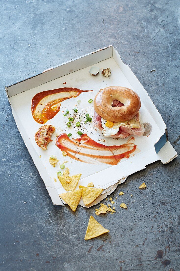 A bagel with egg and bacon in a pizza box with other food remnants