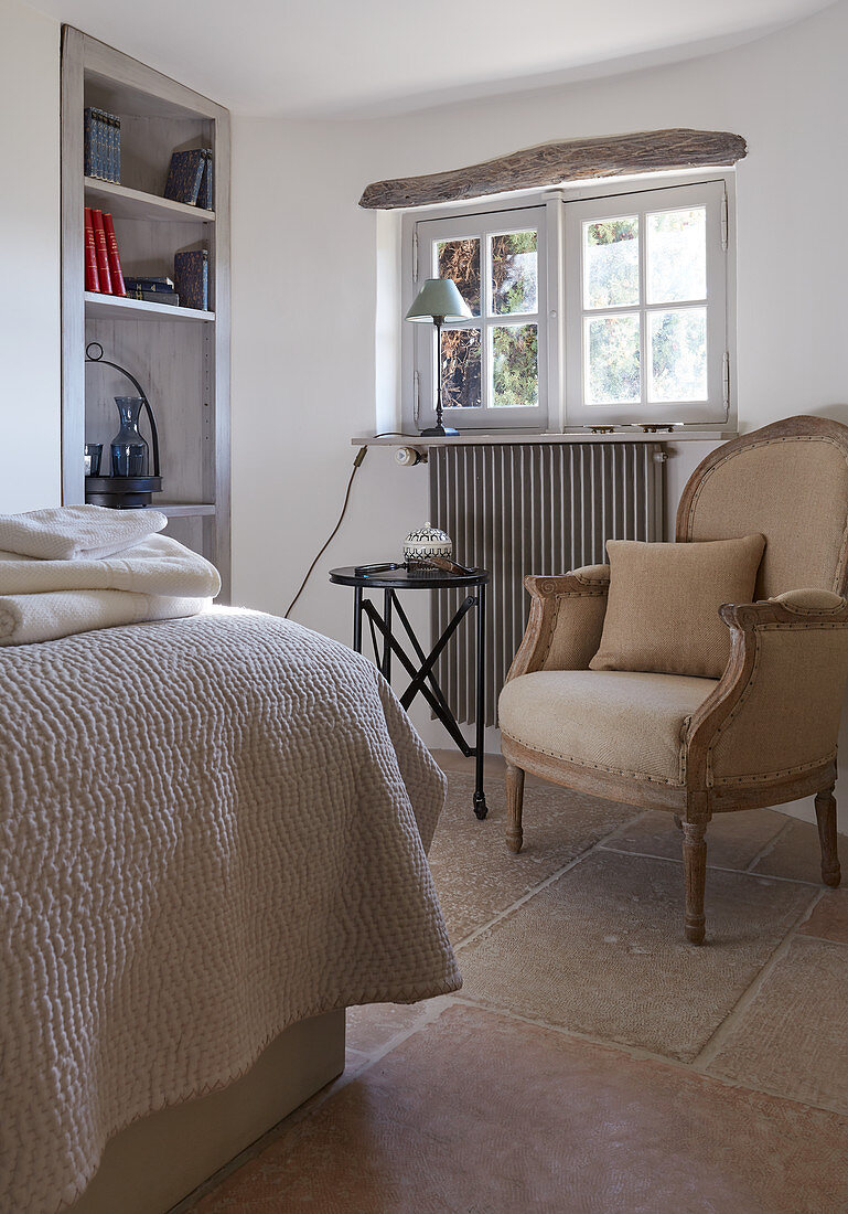 Antique armchair with cushion, side table and fitted shelving in rustic bedroom