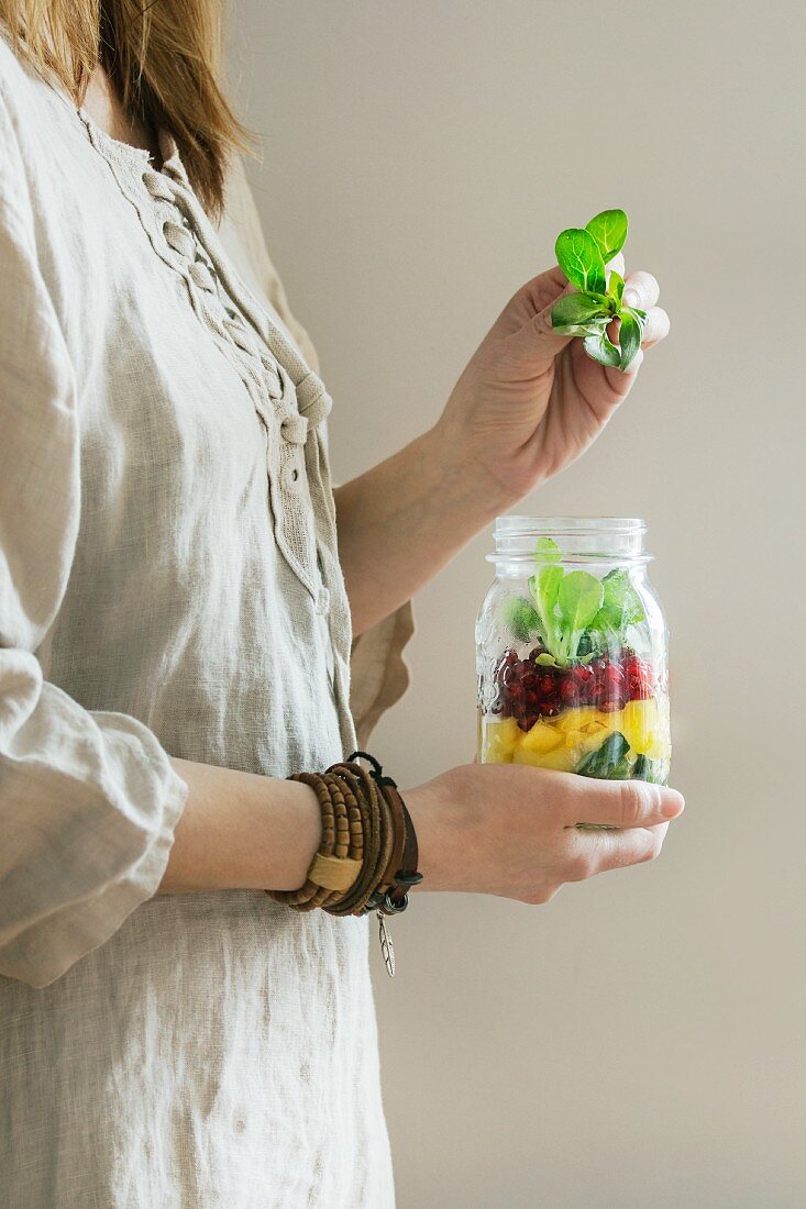 Frau im Leinenkleid hält Einmachglas mit Obstsalat mit Mango, Granatapfelkernen und Feldsalat