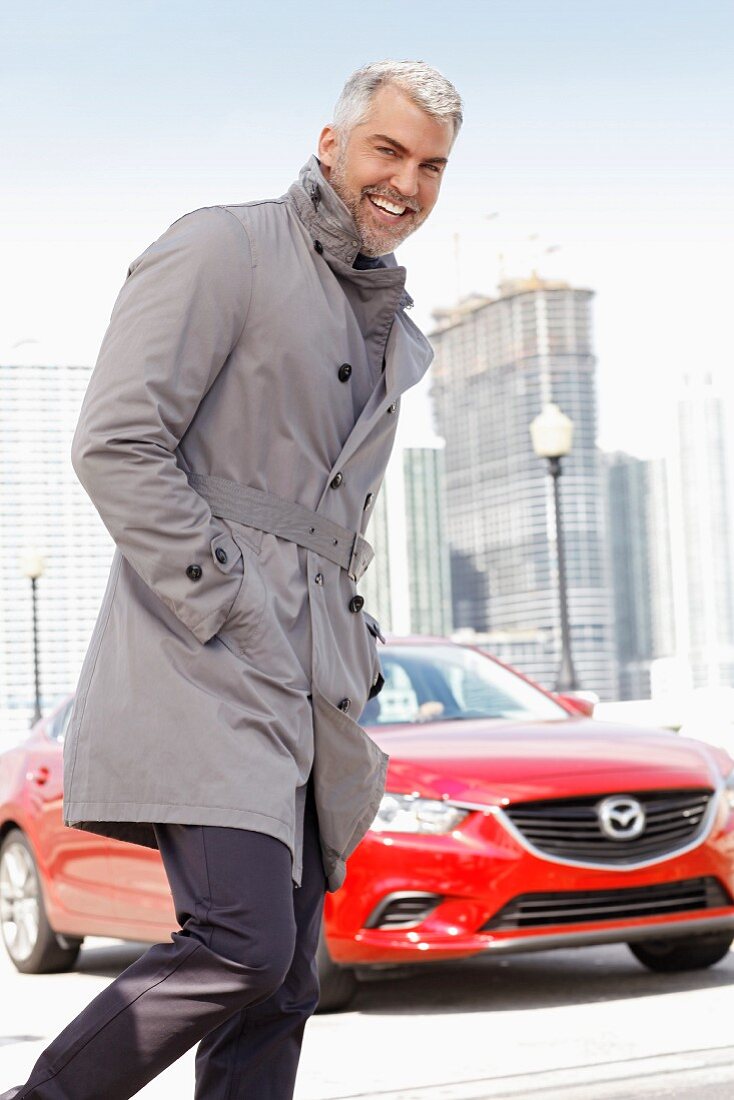 A man with grey hair wearing a grey trenchcoat and trouser suit in front of a red car
