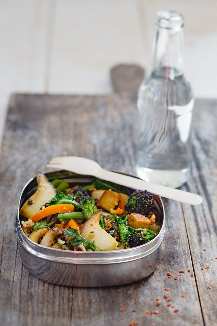 Lentils and rice with mushrooms, carrots and broccoli in a lunch box in front of a water bottle