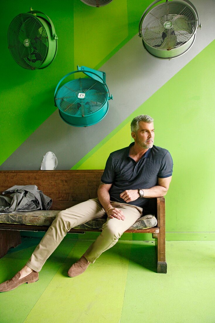 A man with grey hair wearing a T-shirt and chinos sitting on a wooden bench in front of a green background