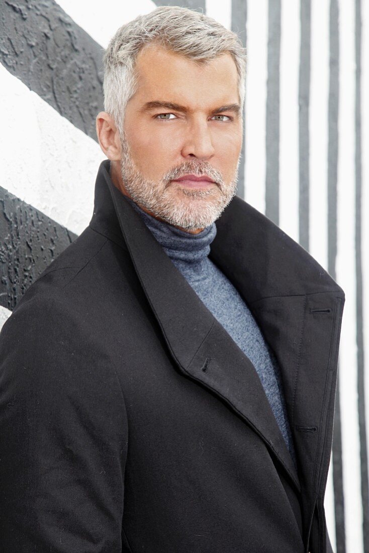 A man with grey hair wearing a roll-neck jumper and a coat in front of a black-and-white wall