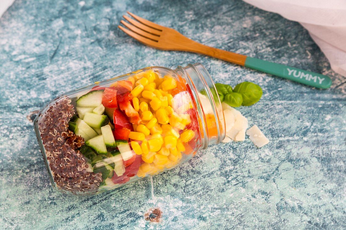 A colourful salad in a glass jar with red quinoa, cucumber, peppers, corn, tomatoes, pecorino and basil, with a wooden fork