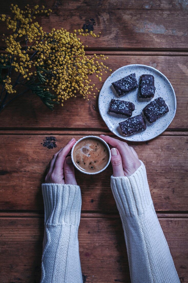 Woman s hands holding a cup of coffee on a rustic wooden table, plate with chocolat coconut bars