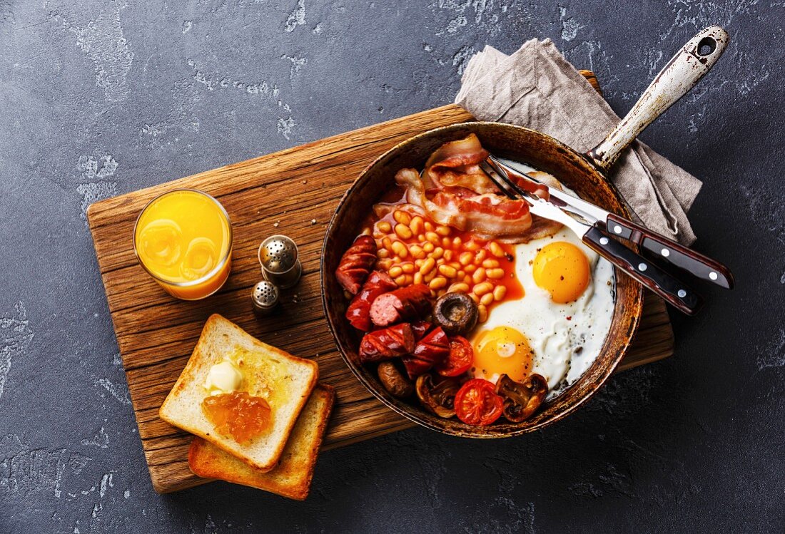 English Breakfast in cooking pan with fried eggs, sausages, bacon, beans, toasts and orange juice on dark stone background