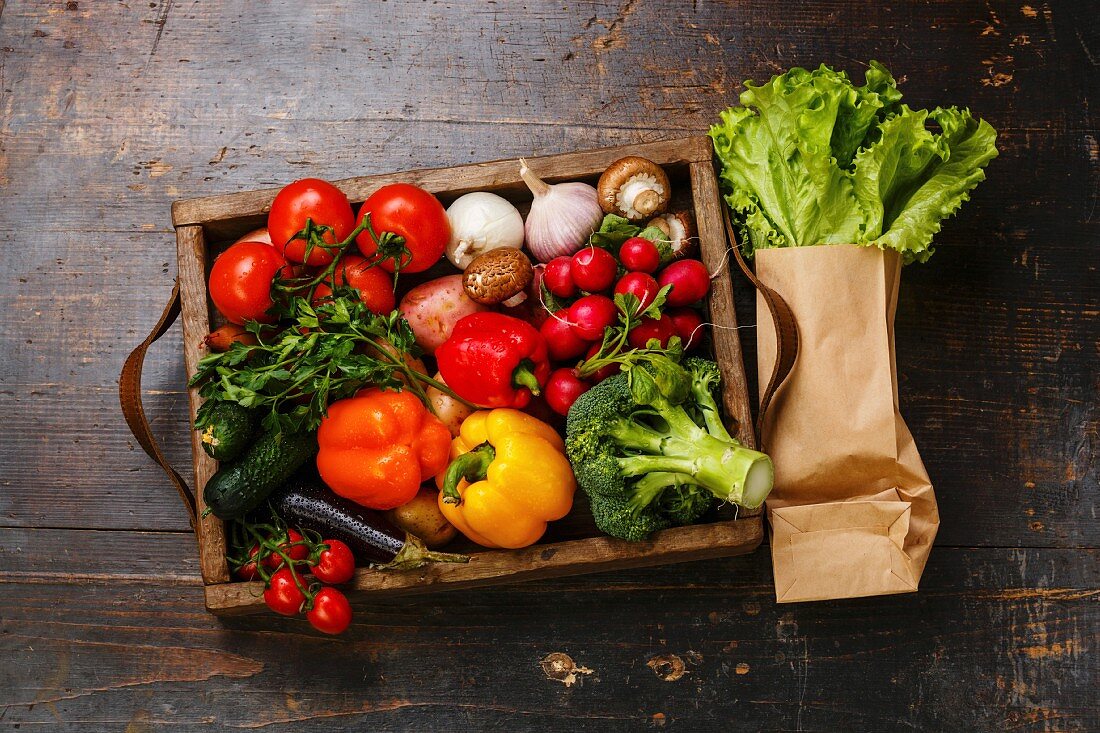 Frisches Gemüse in Holzkiste und Salat in Papiertüte auf Holzuntergrund (Aufsicht)