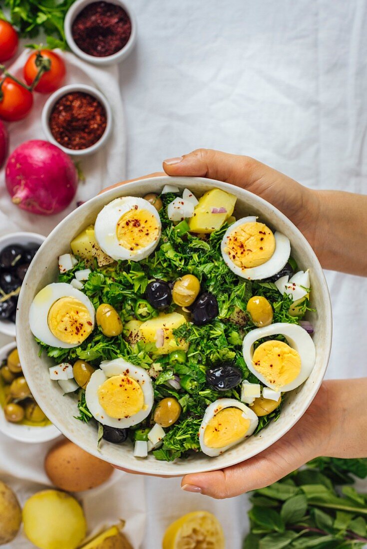Hände halten eine Schüssel Kartoffelsalat mit Kräutern, Eiern und Oliven