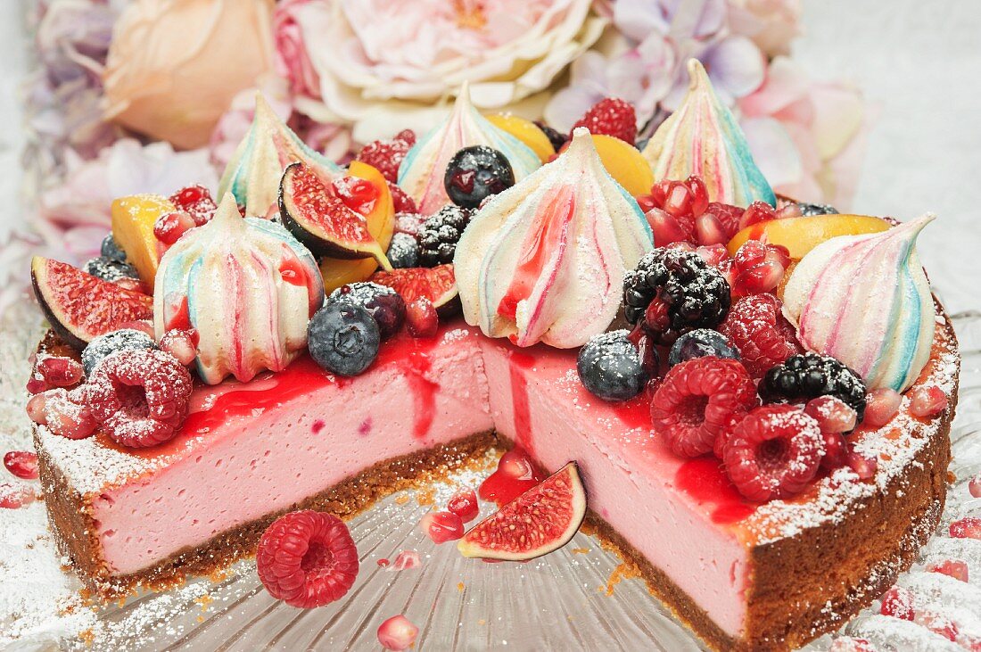 Strawberry cheesecake with merangue kisses and fresh fruit on the top on a glass cake stand in front of flowers