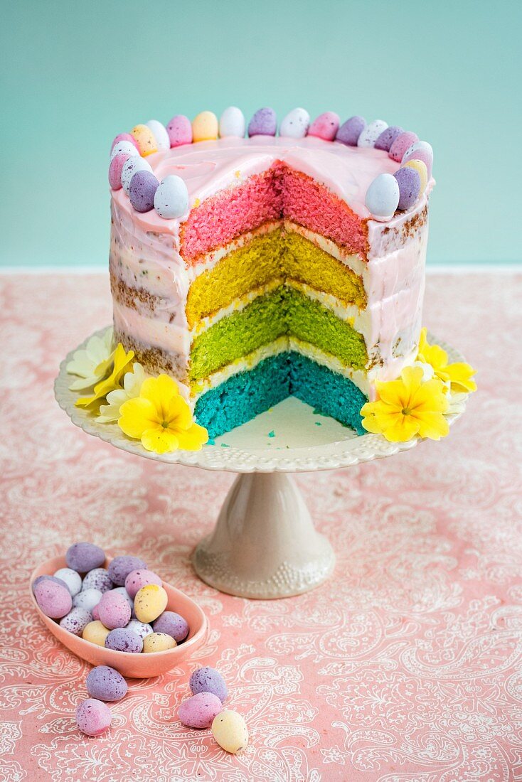 A rainbow cake decorated with mini chocolate eggs and flowers for Easter (with a slice cut out)