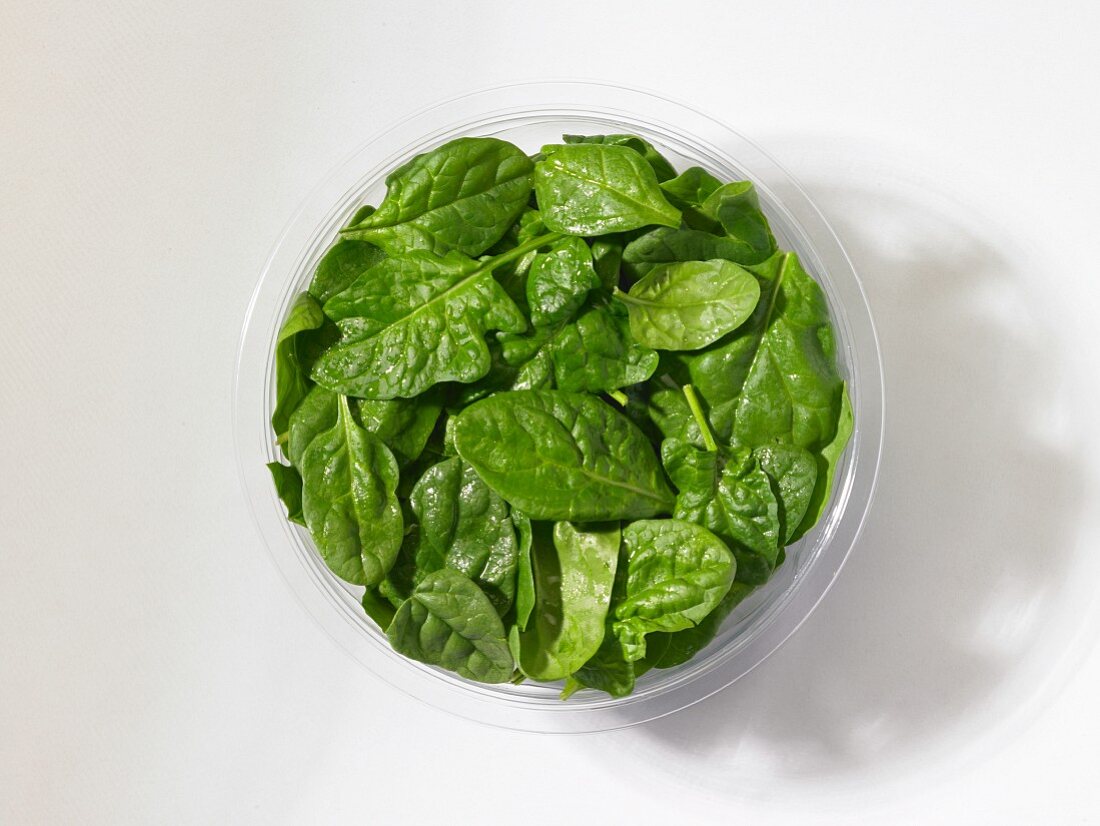 Fresh baby spinach in a plastic bowl in front of a white background (seen from above)