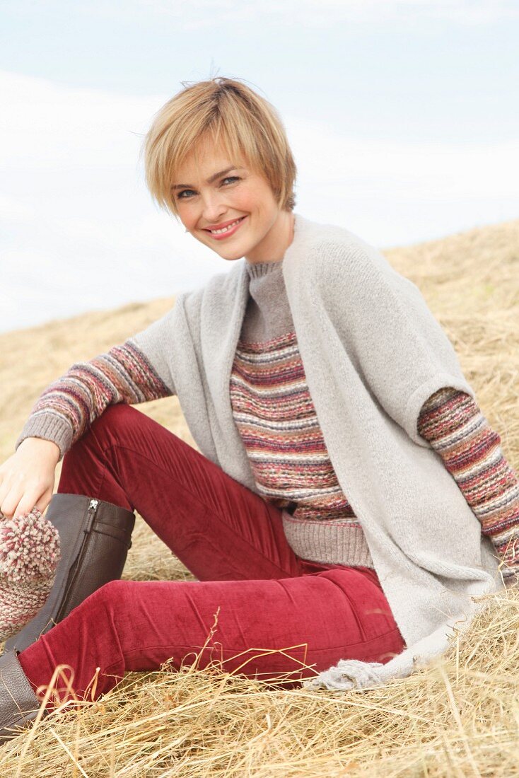 A blonde woman wearing a striped knitted jumper, a poncho, red velvet trousers and boots holding a knitted hat