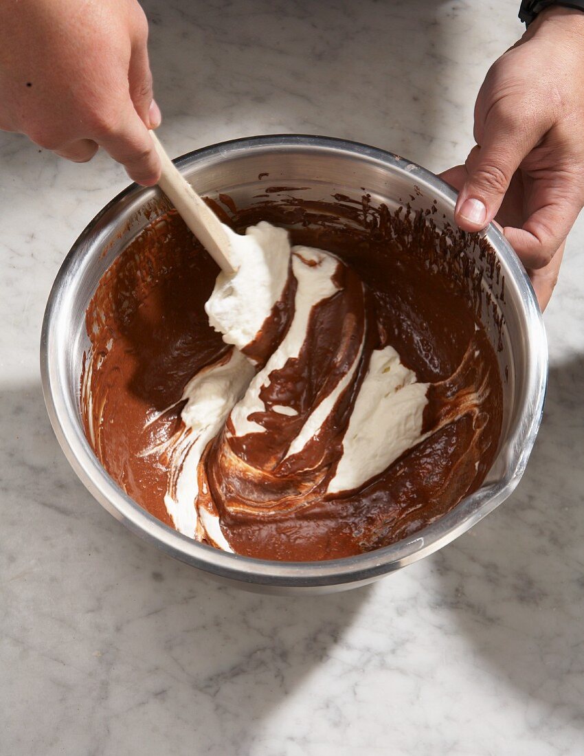 Cream being mixed into melted chocolate with a dough scraper in a metal bowl