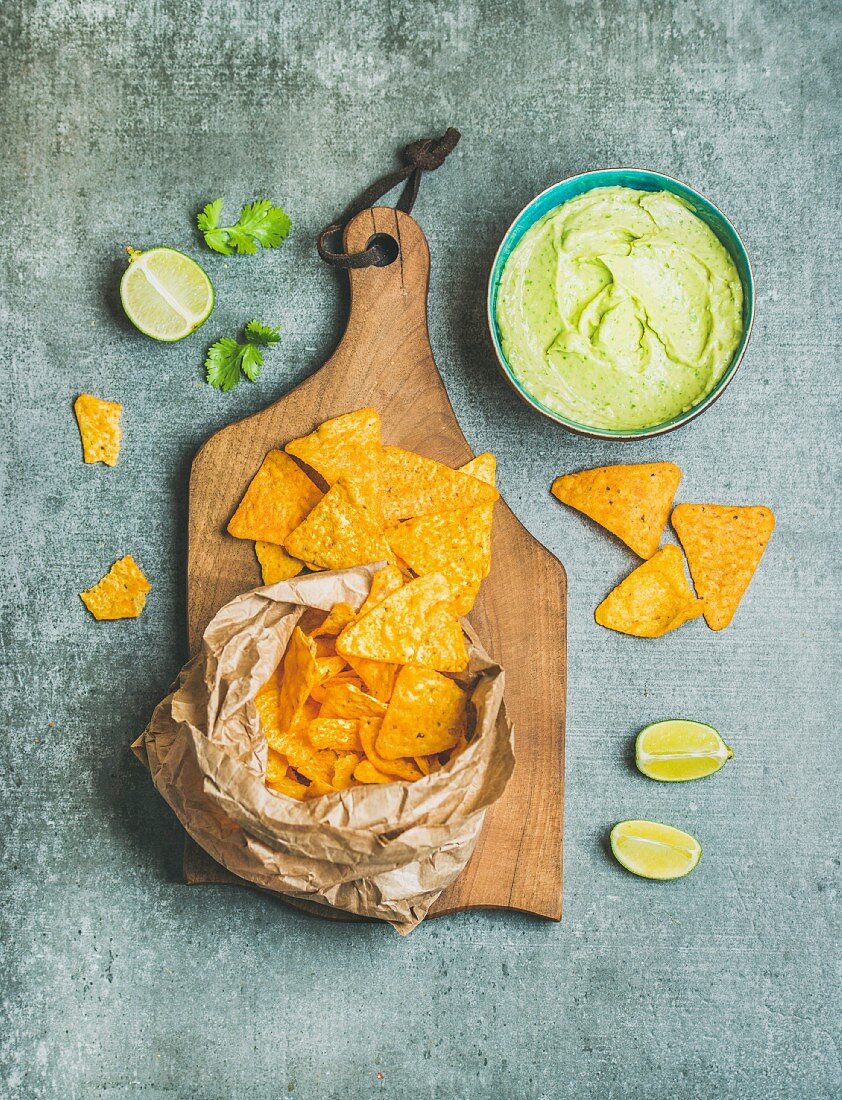 Mexican corn chips and fresh guacamole sauce on wooden serving board over grey concrete table background, top view, vertical composition