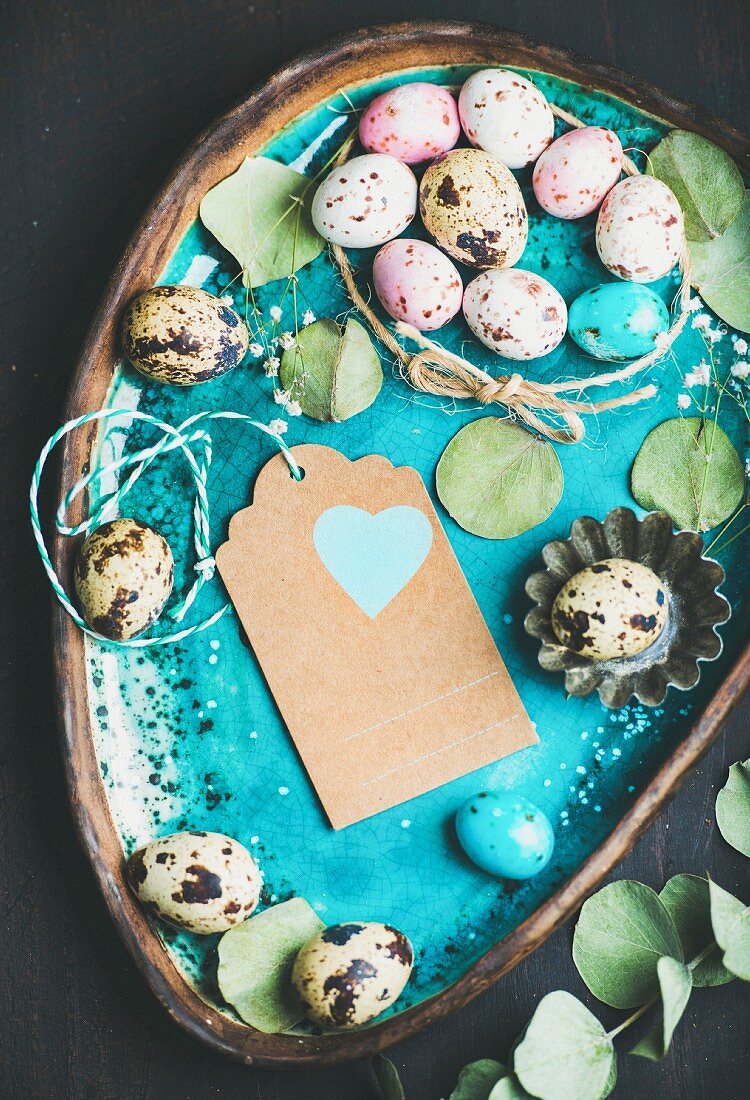 Colorful quail eggs, dried flowers and leaves for Easter holiday over turquoise blue tray with craft paper label