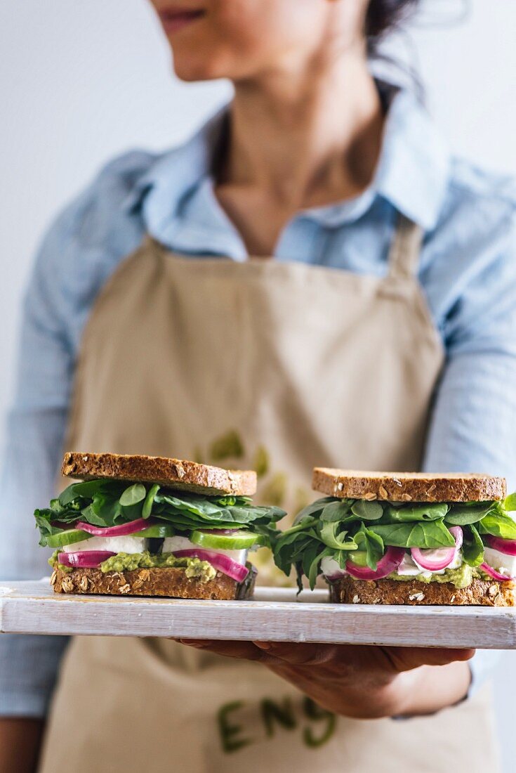 A woman with a blue shirt and a beige apron holding springtime feta sandwiches with herbs, avocado and pickled red onions