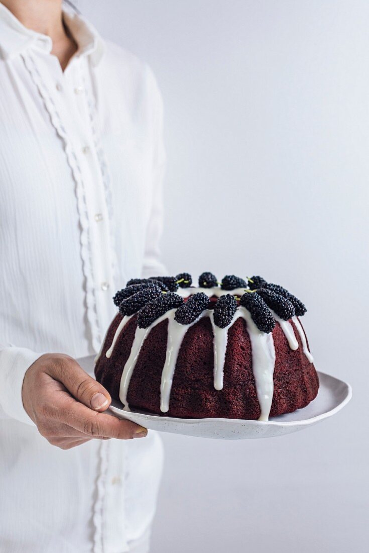 A woman holding a red velvet bundt cake with a white glaze and black mulberries on the top