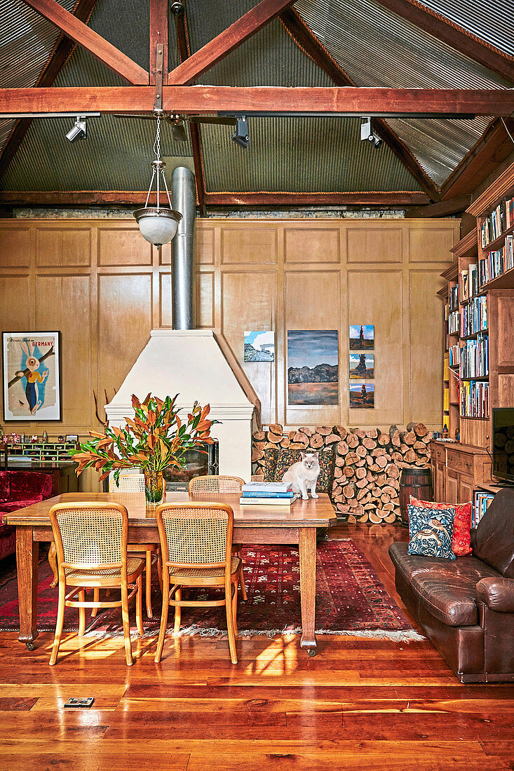 Dining table with chairs, vintage leather sofa and bookcase in a high living room with fireplace and wood storage on paneling